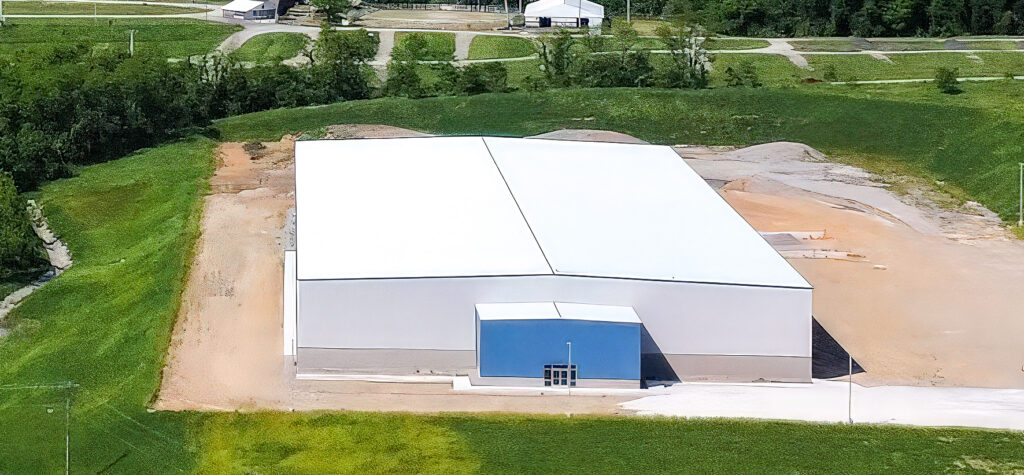 Aerial view of a large white industrial building in Dunbar with a blue entrance, surrounded by paved areas, green fields, and additional buildings in the background